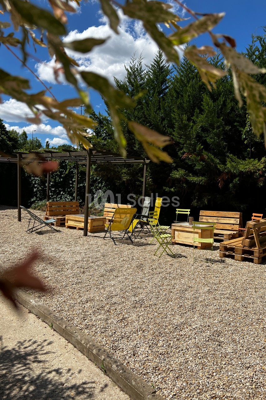 Espace extérieur avec chaises colorées et mobilier en palettes sur un sol gravillonné, entouré de végétation.