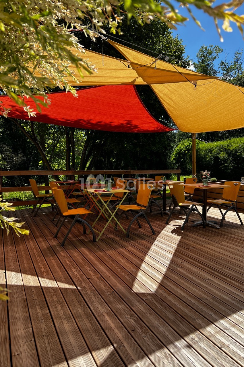 Terrasse en bois avec des tables et chaises jaunes, ombragée par des voiles rouges et jaunes, entourée de verdure.