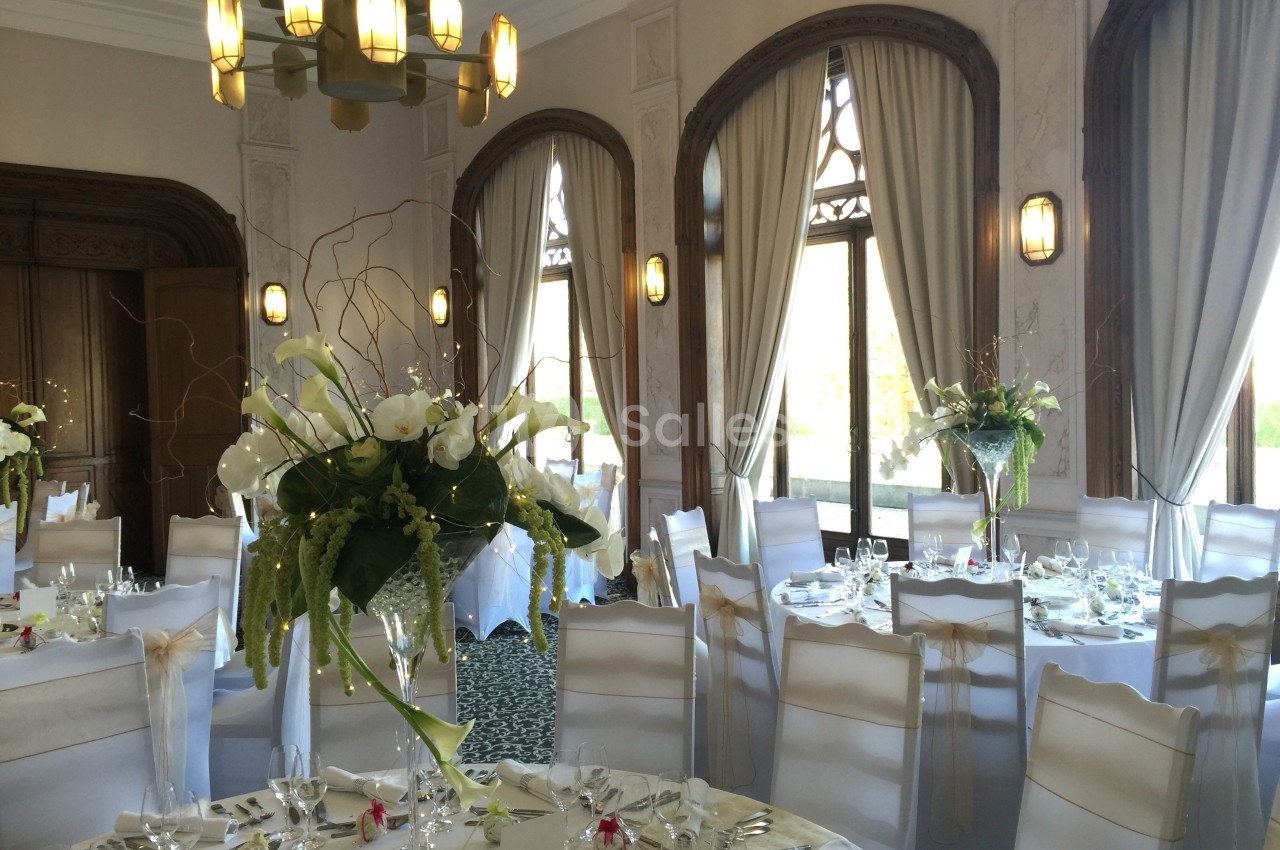 Salle de réception élégante avec tables rondes dressées, chaises blanches et grandes compositions florales.