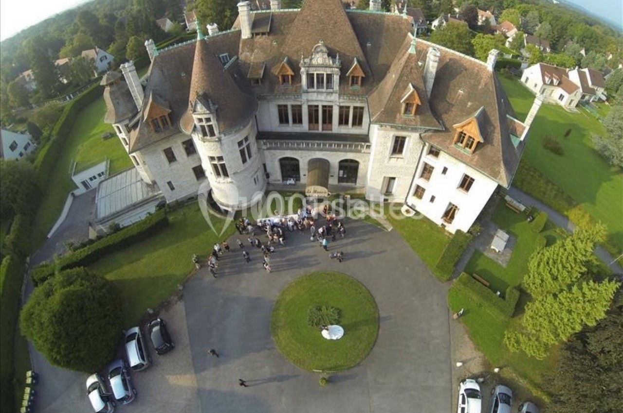 Vue aérienne d'un grand manoir avec une cour centrale où des personnes sont rassemblées, entouré de verdure.