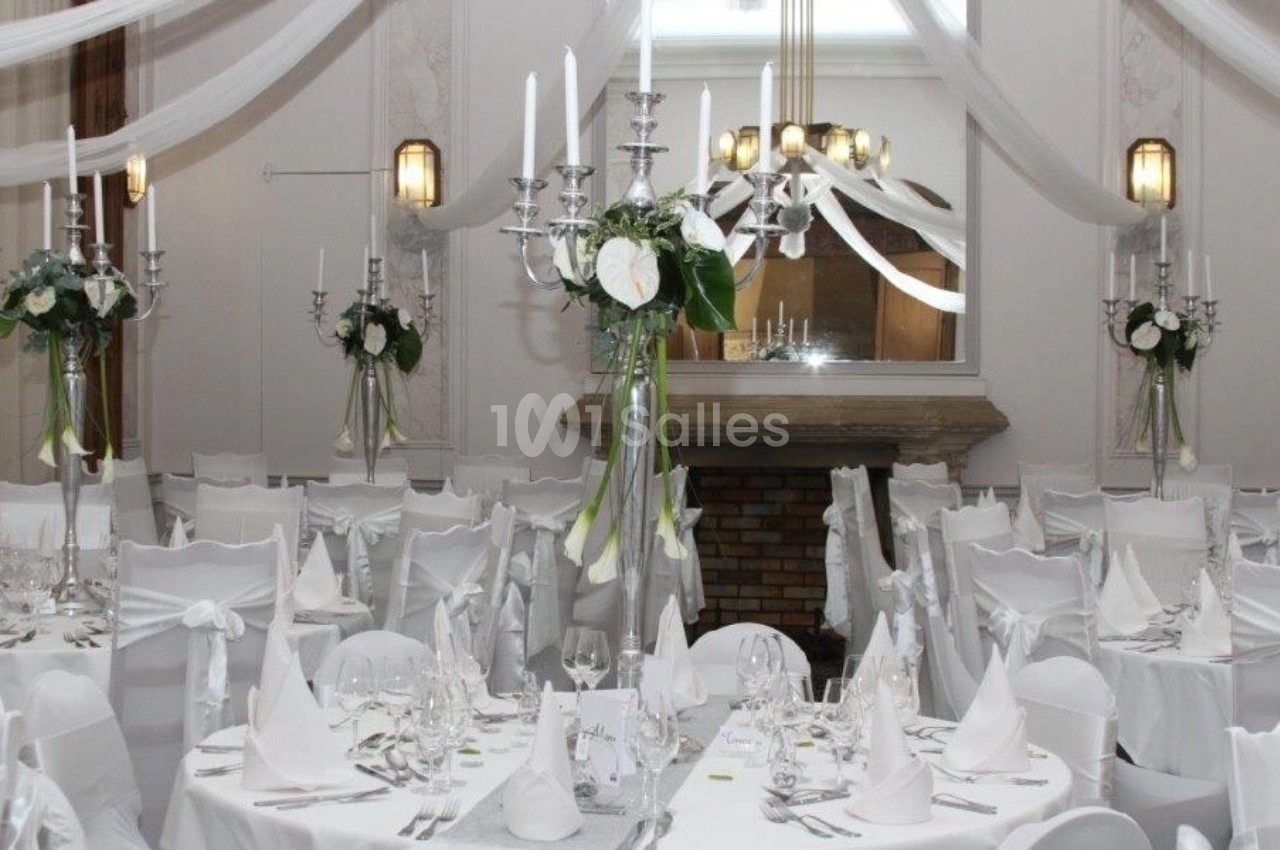 Salle de réception décorée en blanc avec chandeliers, nappes élégantes et compositions florales sur les tables.