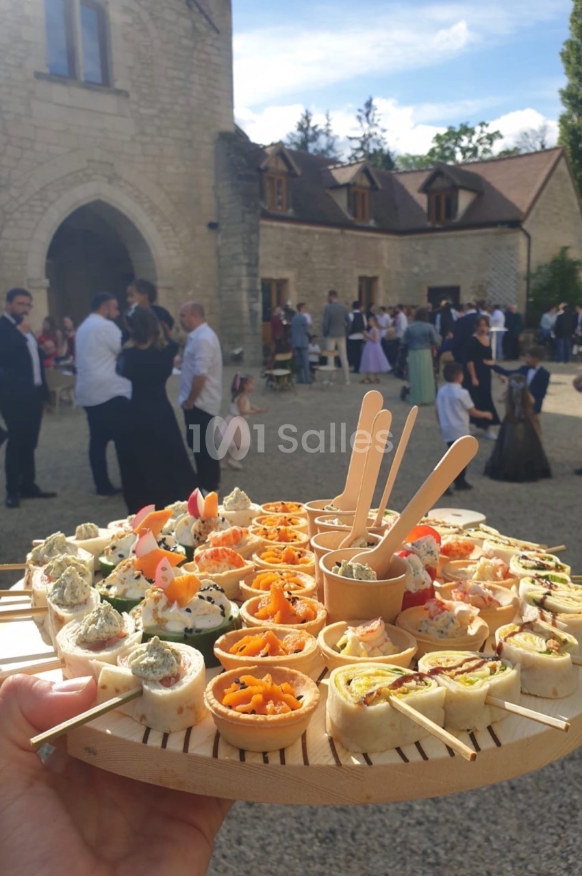 Plateau de bouchées apéritives variées tenu en main, avec des invités rassemblés devant un bâtiment en pierre.