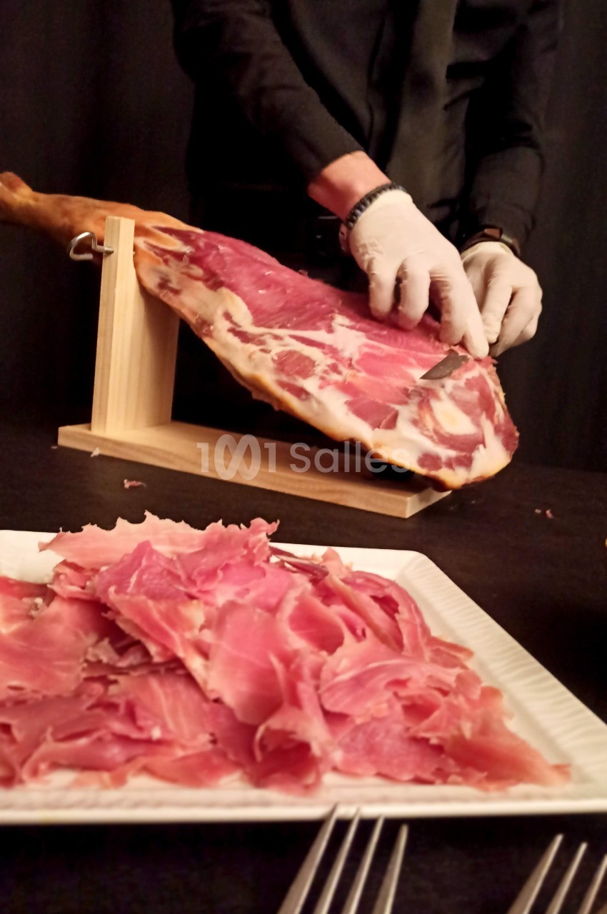 Un serveur découpe finement un jambon sur un support en bois, avec des tranches disposées sur une assiette au premier plan.