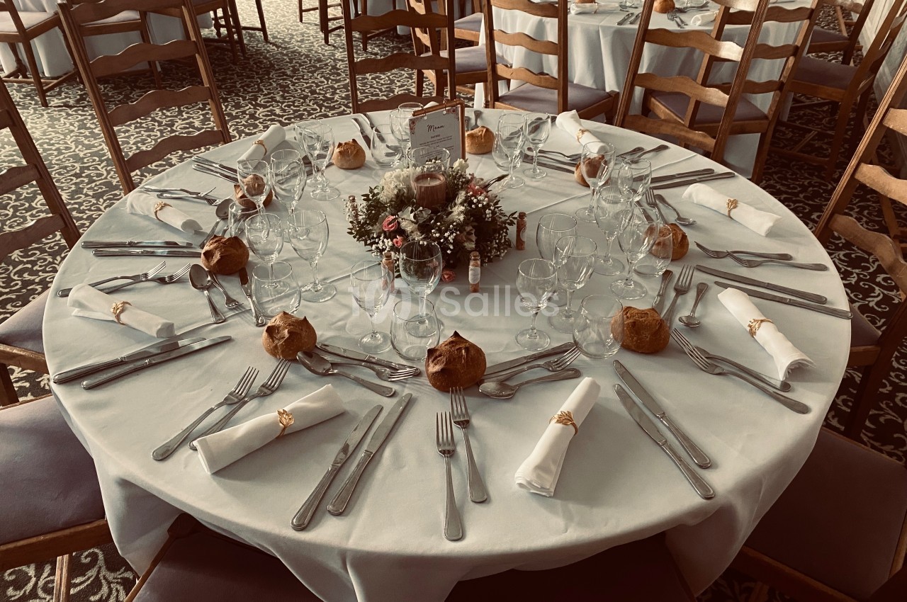 Table ronde dressée pour un repas, avec nappes blanches, couverts, verres, serviettes et décoration florale centrale.