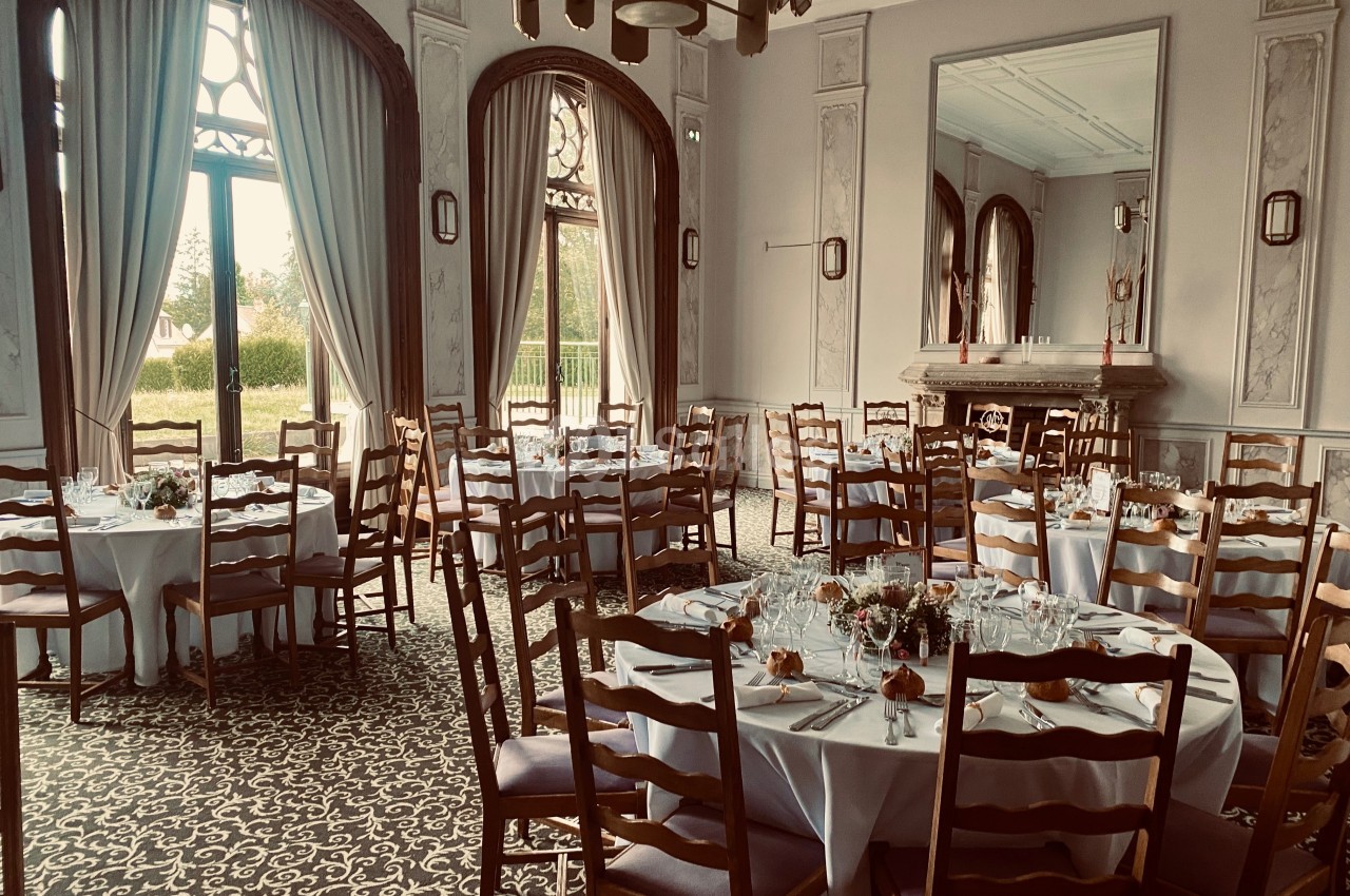 Salle élégante avec tables rondes dressées, chaises en bois, grandes fenêtres et cheminée décorative en arrière-plan.