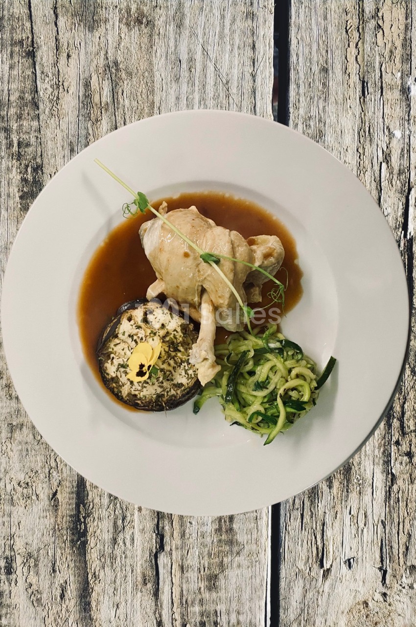 Assiette avec un suprême de volaille, une courgette spiralée, un champignon farci et une sauce brune, sur une table en bois.
