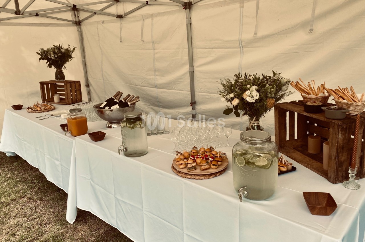 Buffet sous une tente avec boissons, verrines, pains et décorations florales sur une table blanche.