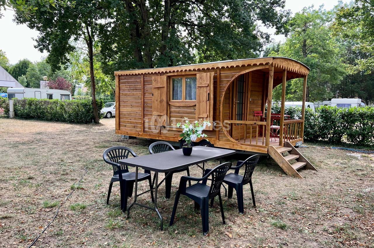 Roulotte en bois installée sur un terrain herbeux avec une table et des chaises en plastique noir à l'extérieur.