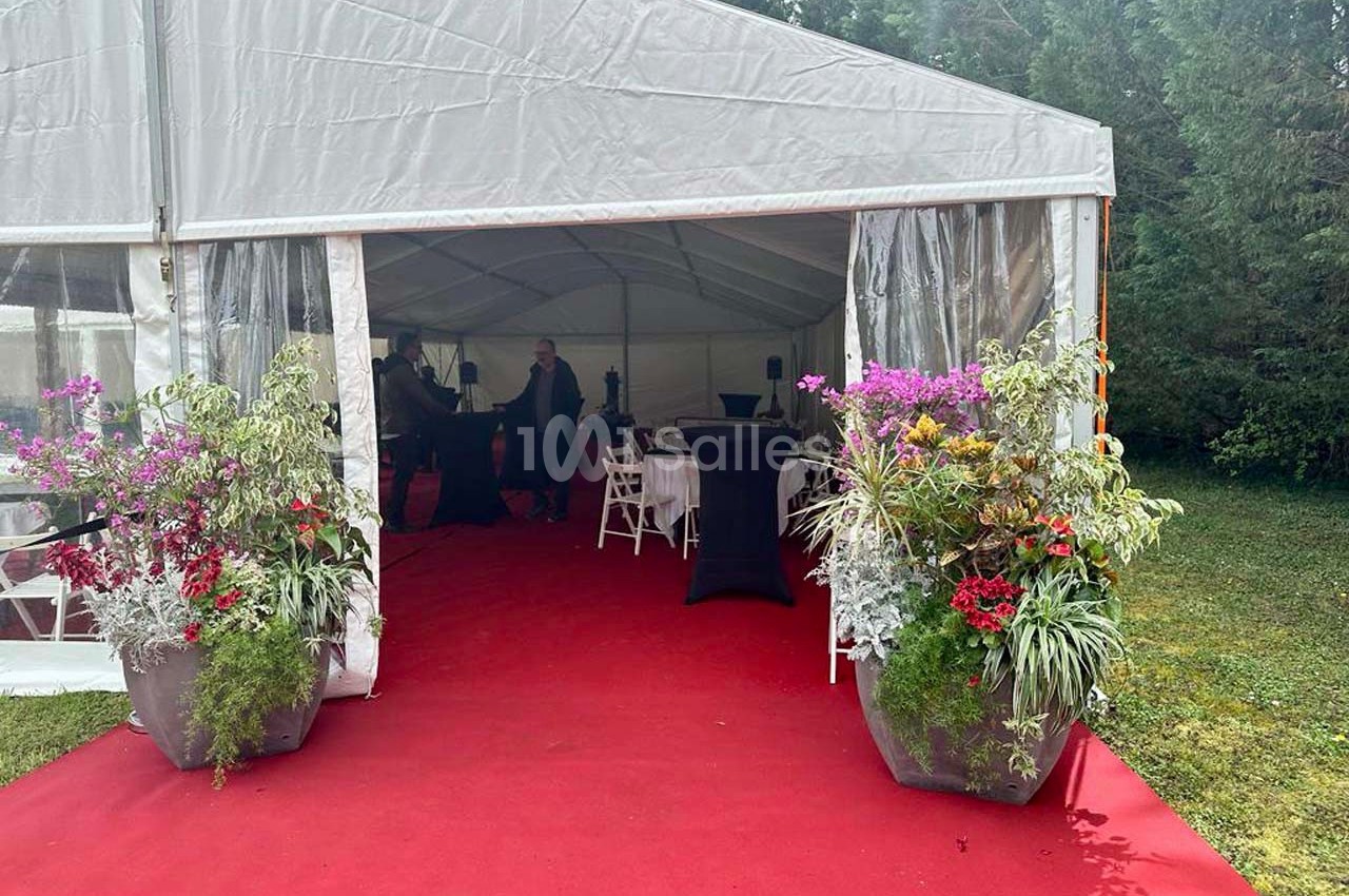 Entrée d'une tente événementielle avec tapis rouge, grandes plantes fleuries et mobilier de réception à l'intérieur.