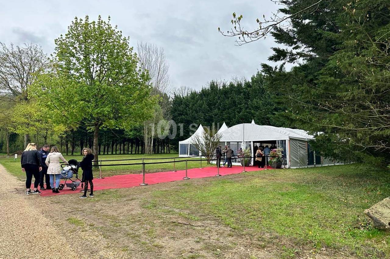 Tente blanche avec tapis rouge menant à l'entrée, entourée d'arbres, avec des visiteurs arrivant.