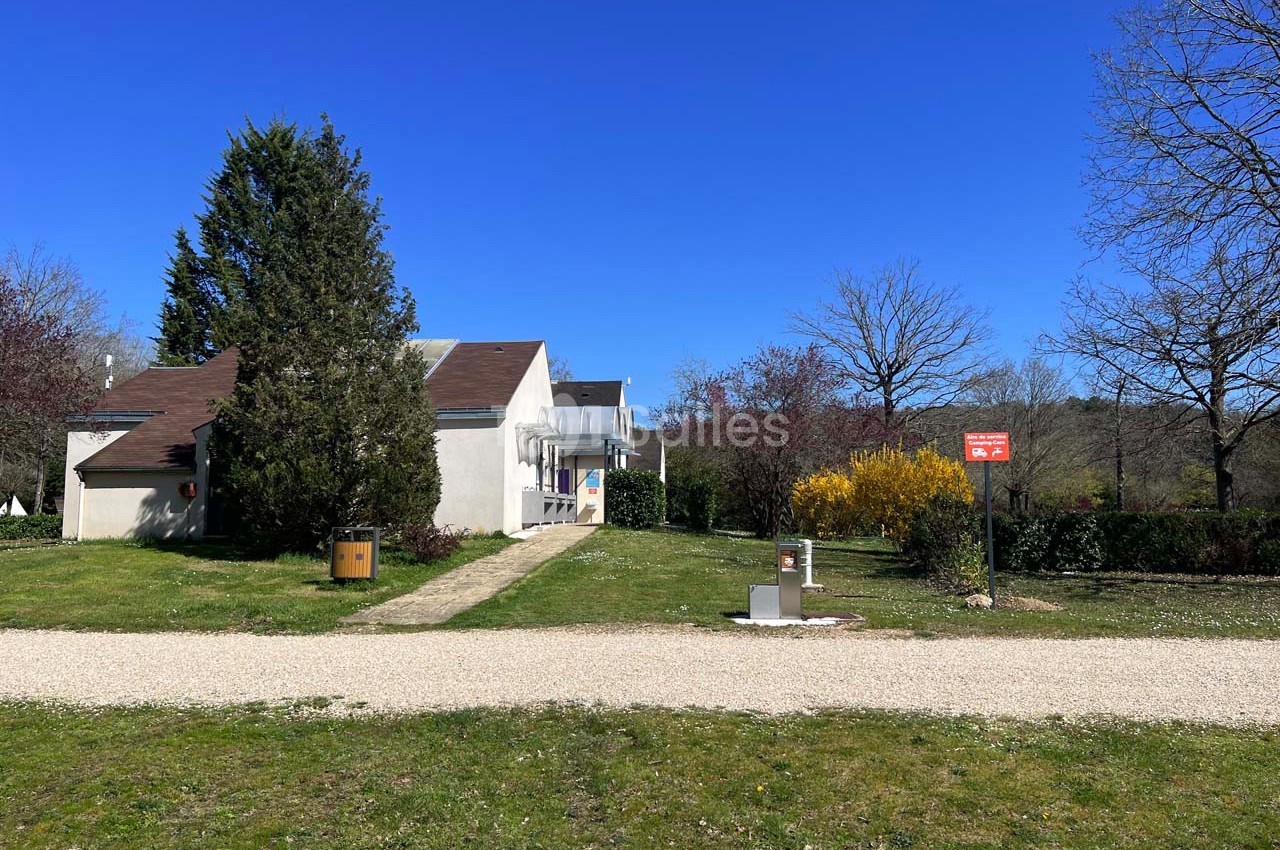 Maison blanche avec toit brun entourée de végétation, chemin en gravier et panneau rouge visible au premier plan.