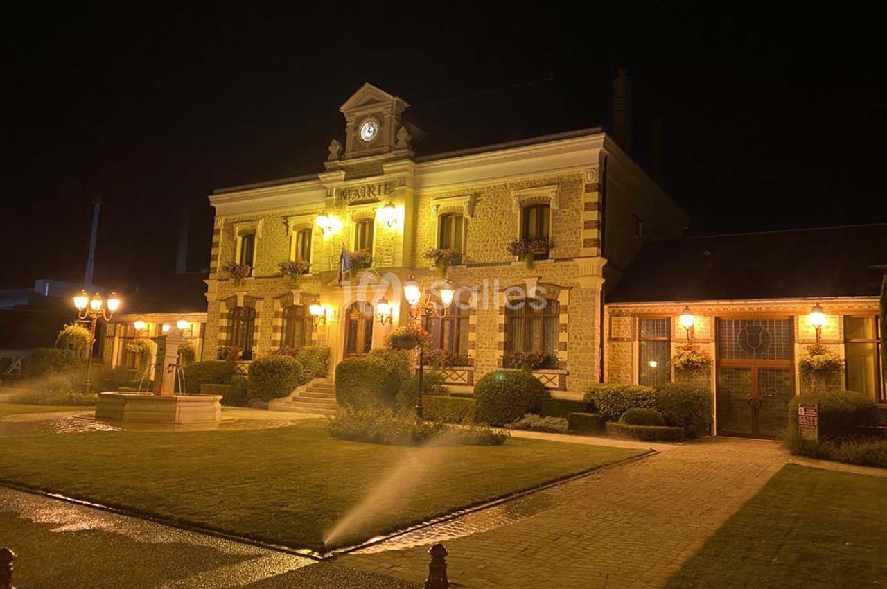 Bâtiment éclairé de nuit avec une horloge en façade, entouré de jardins et d'allées arrosées.