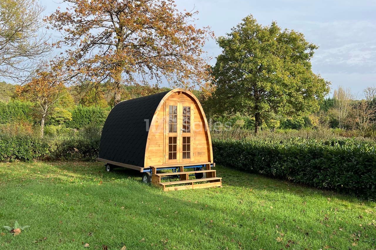 Cabane en bois de forme arrondie avec toit sombre, située sur une pelouse entourée d'arbres et de haies.