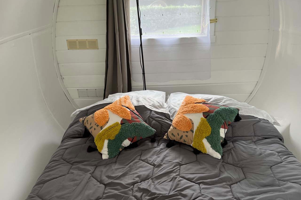 Lit double avec couette grise, deux coussins colorés et une fenêtre avec rideaux dans une pièce blanche.