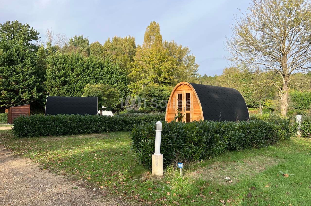 Deux cabanes en bois en forme d'arc entourées de haies, situées dans un espace vert arboré.