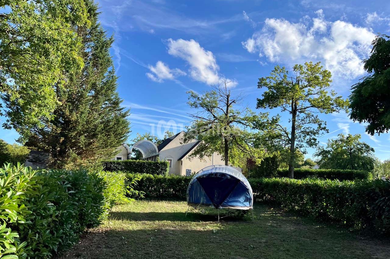 Tente bleue installée sur une pelouse entourée de haies et d'arbres, avec une maison blanche en arrière-plan.
