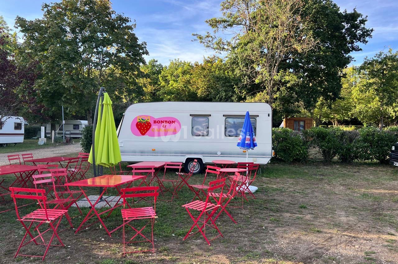 Caravane blanche avec un logo de fraise, entourée de tables et chaises rouges dans un espace extérieur arboré.