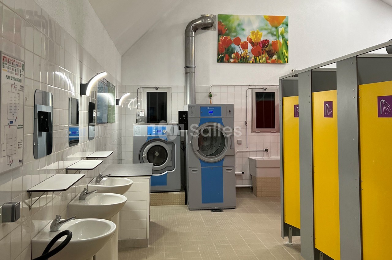Laverie avec machines à laver, sèche-linge, éviers, miroirs et cabines jaunes dans un espace carrelé.
