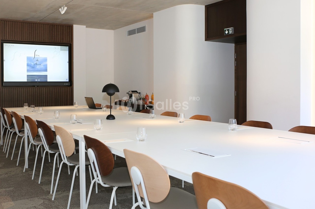 Salle de réunion moderne avec une longue table blanche, des chaises en bois, un écran allumé et un coin rafraîchissements.