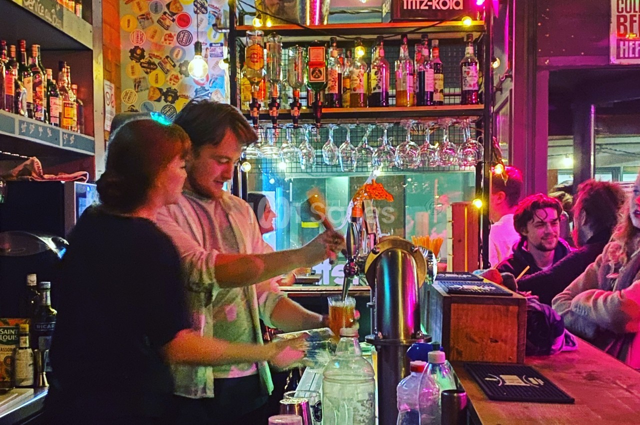 Un barman et une barmaid préparent des boissons dans un bar animé, entourés de bouteilles et de lumières colorées.