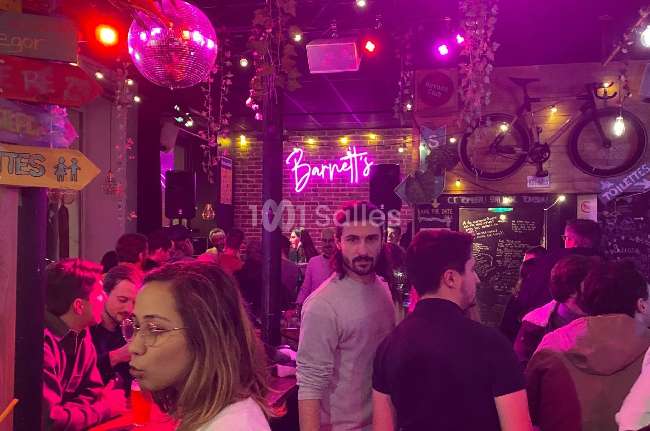 Bar animé avec des lumières colorées, des clients discutant et un décor comprenant un vélo et un néon ’Barretts’.