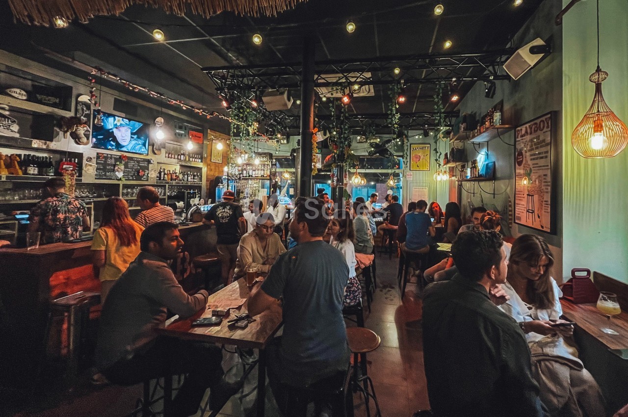Personnes assises et debout dans un bar animé, éclairé par des lumières chaleureuses et décoré de plantes.