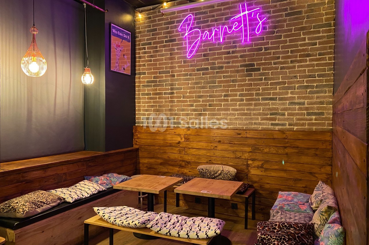 Espace cosy avec banquettes, coussins colorés, murs en briques, éclairage doux et enseigne néon violette.