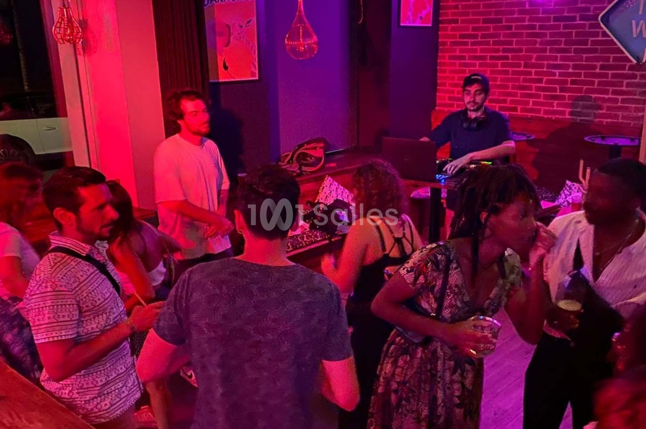 Des personnes dansent et discutent dans un bar à l'éclairage rouge, avec un DJ en arrière-plan.