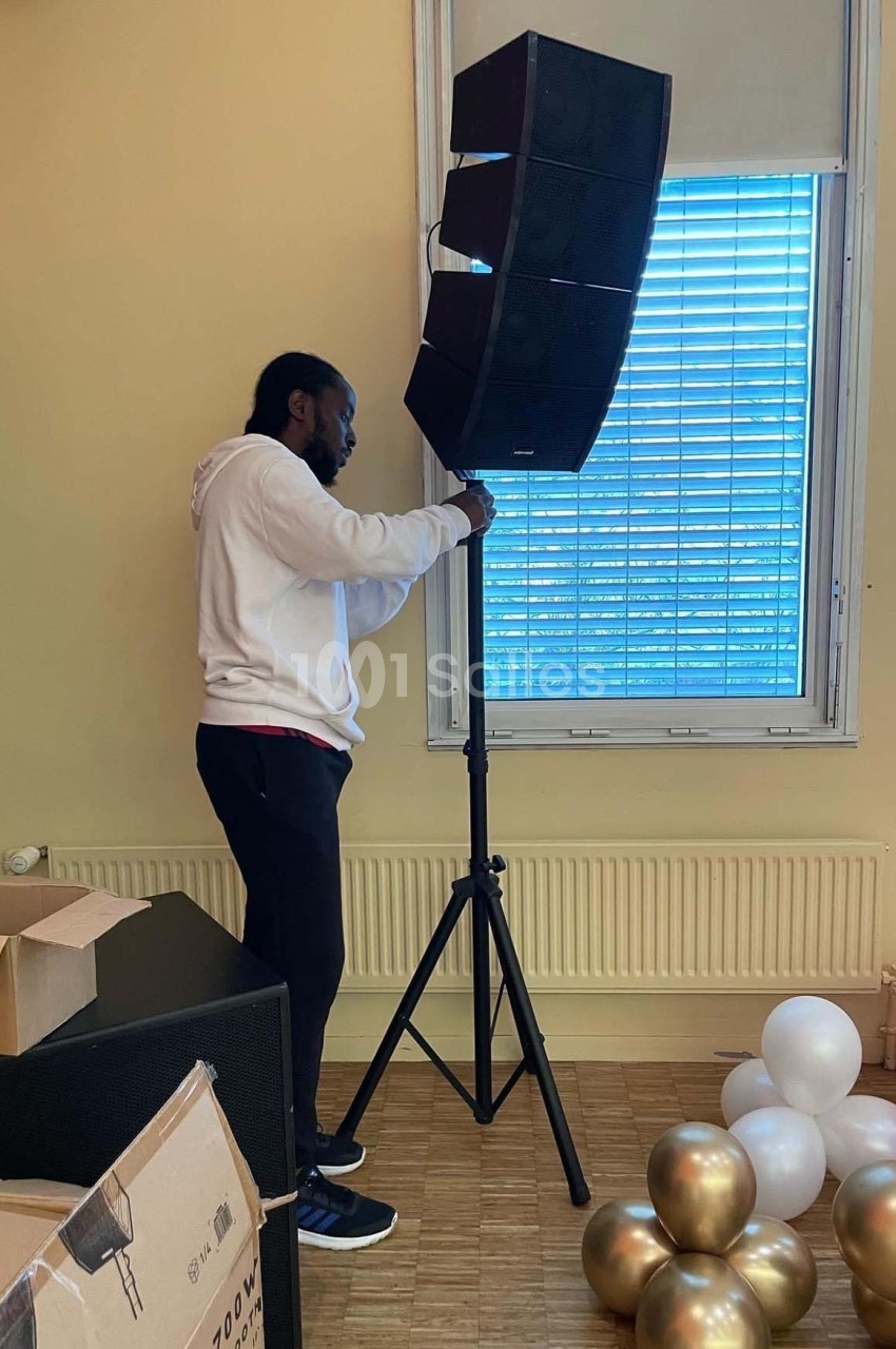 Un homme ajuste un haut-parleur sur un trépied dans une pièce avec des ballons et des cartons au sol.