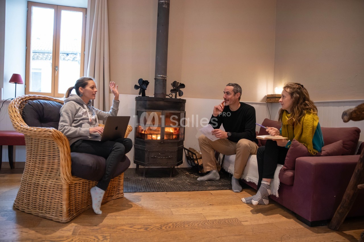 Trois personnes discutent dans un salon chaleureux près d'un poêle à bois, avec des fauteuils et une lumière naturelle.