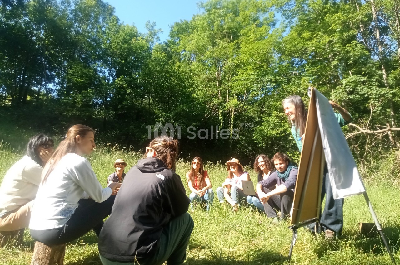 Un groupe de personnes assises en cercle dans un pré, écoutant une présentation devant un tableau chevalet.