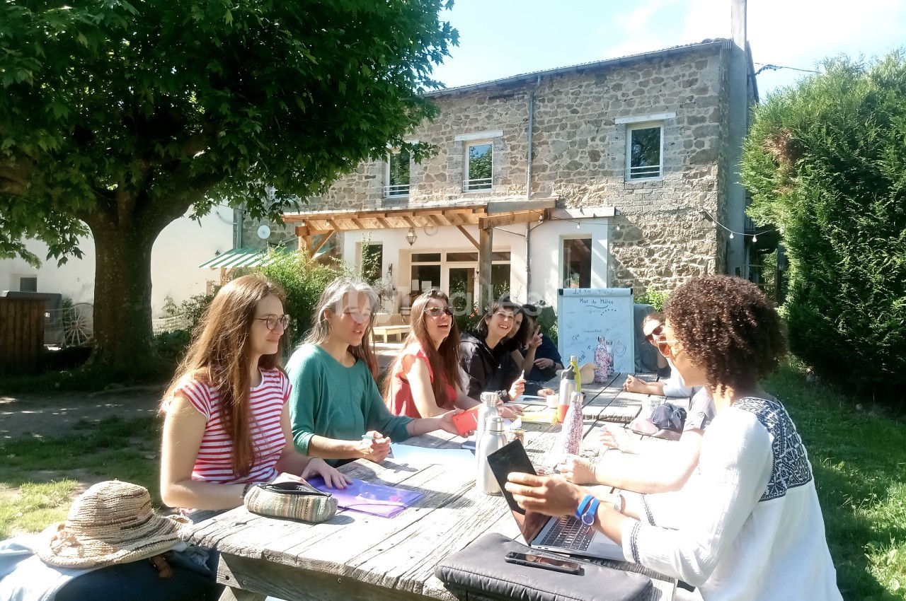 Un groupe de personnes assises autour d'une table en bois dans un jardin, discutant devant une maison en pierre.