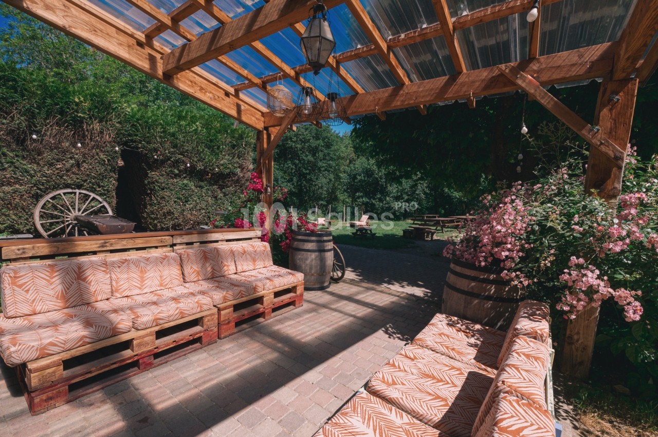 Terrasse en bois avec banquettes à motifs, fleurs roses, barriques et vue sur un jardin verdoyant en arrière-plan.