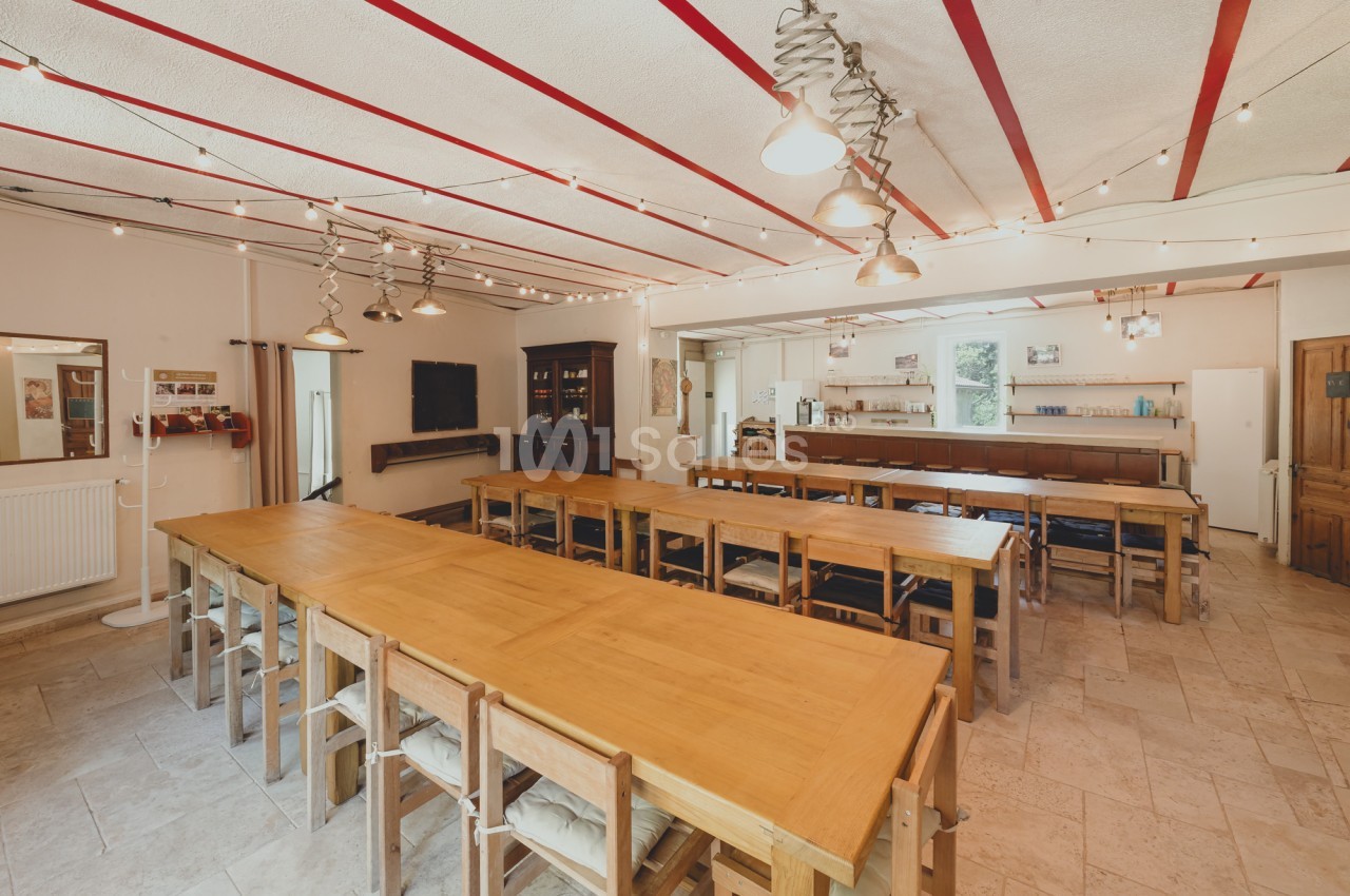 Salle lumineuse avec de longues tables en bois, des chaises assorties et des guirlandes lumineuses au plafond.