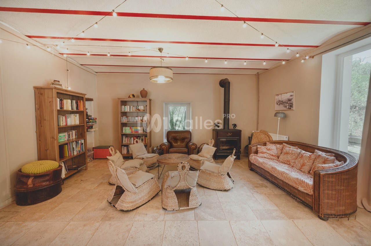 Salon chaleureux avec canapé en rotin, fauteuils en osier, bibliothèque, poêle à bois et guirlande lumineuse au plafond.