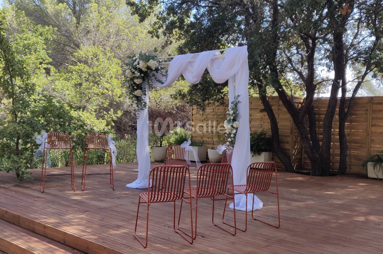 Arche de mariage décorée de voilages et de fleurs blanches sur une terrasse en bois entourée de végétation.
