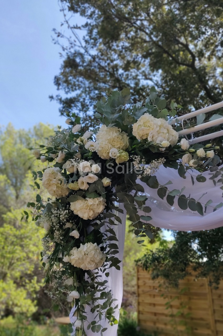 Arrangement floral blanc et vert avec des roses et hortensias décorant une arche en bois sous un ciel dégagé.