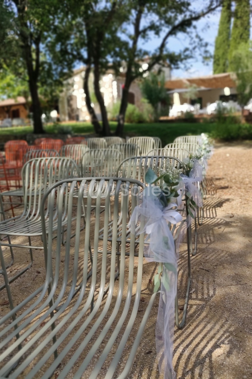 Chaises en métal alignées en extérieur, décorées de rubans blancs, dans un cadre arboré et ensoleillé.