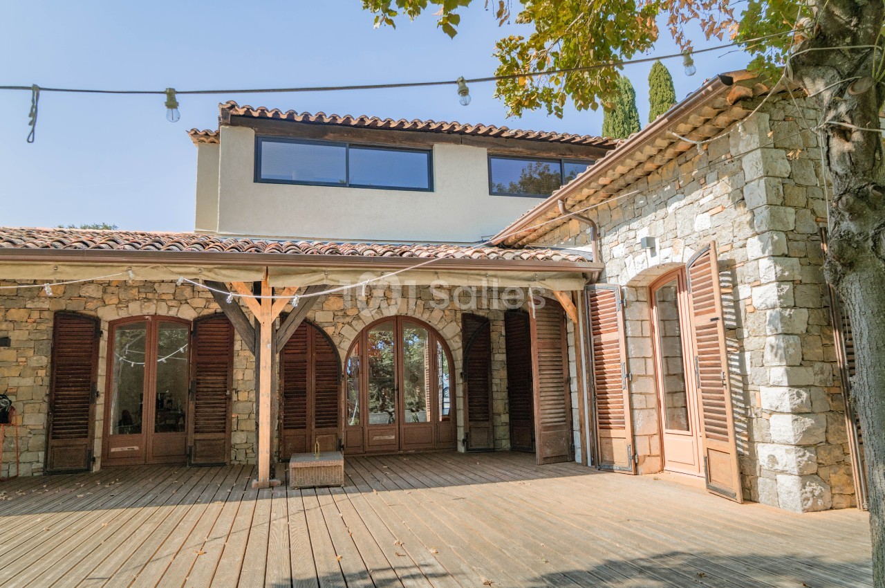 Maison en pierre avec volets en bois, terrasse en bois et toit en tuiles, entourée de végétation.