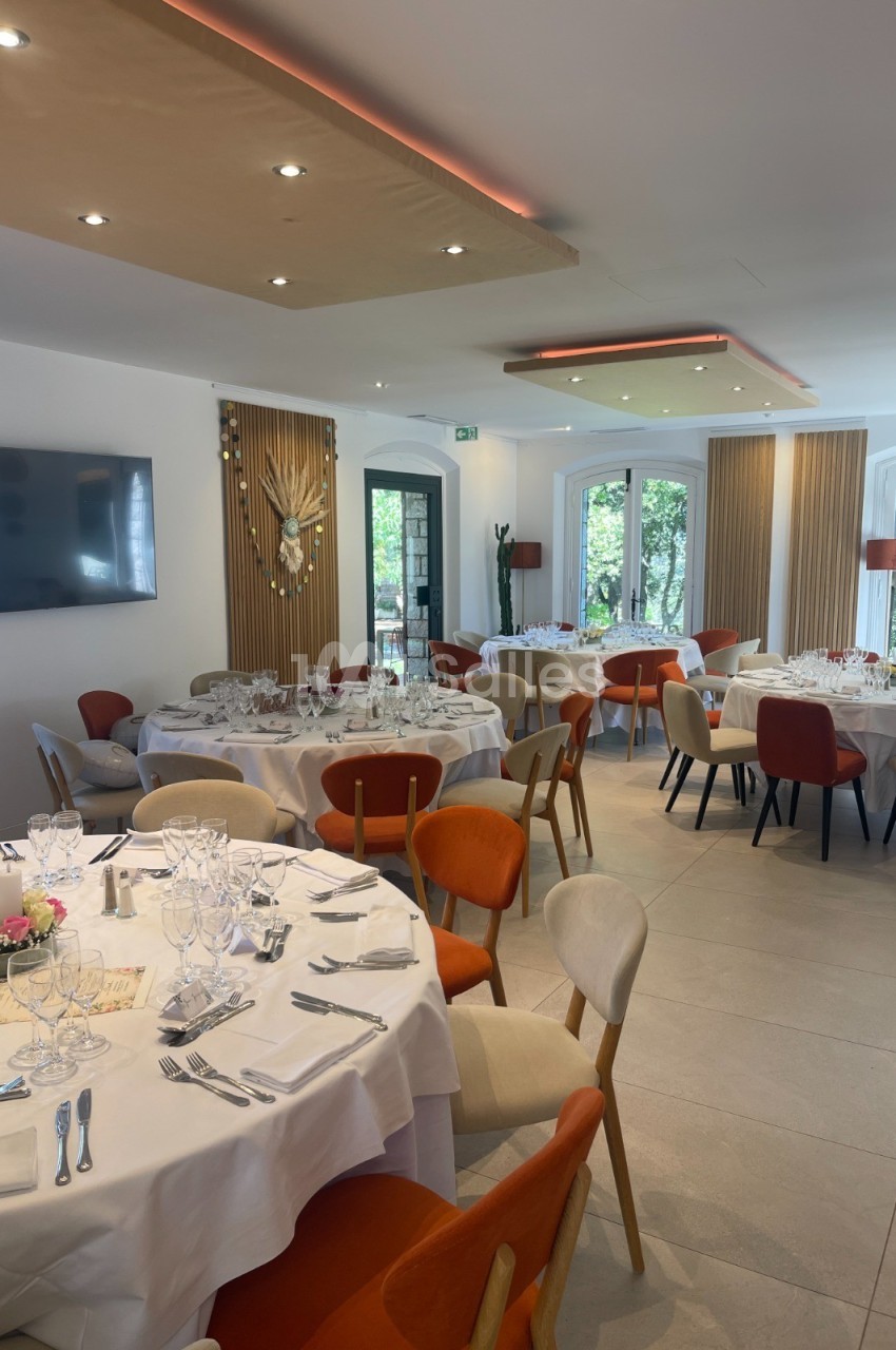 Salle de réception lumineuse avec tables rondes dressées, chaises colorées et grandes baies vitrées donnant sur un jardin.