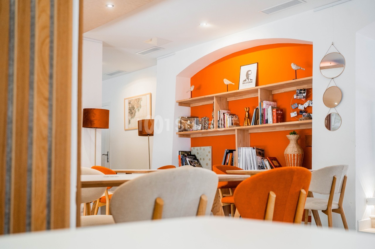 Intérieur contemporain avec mur orange arqué, étagères en bois, chaises beige et orange, mobilier clair et miroirs ronds.