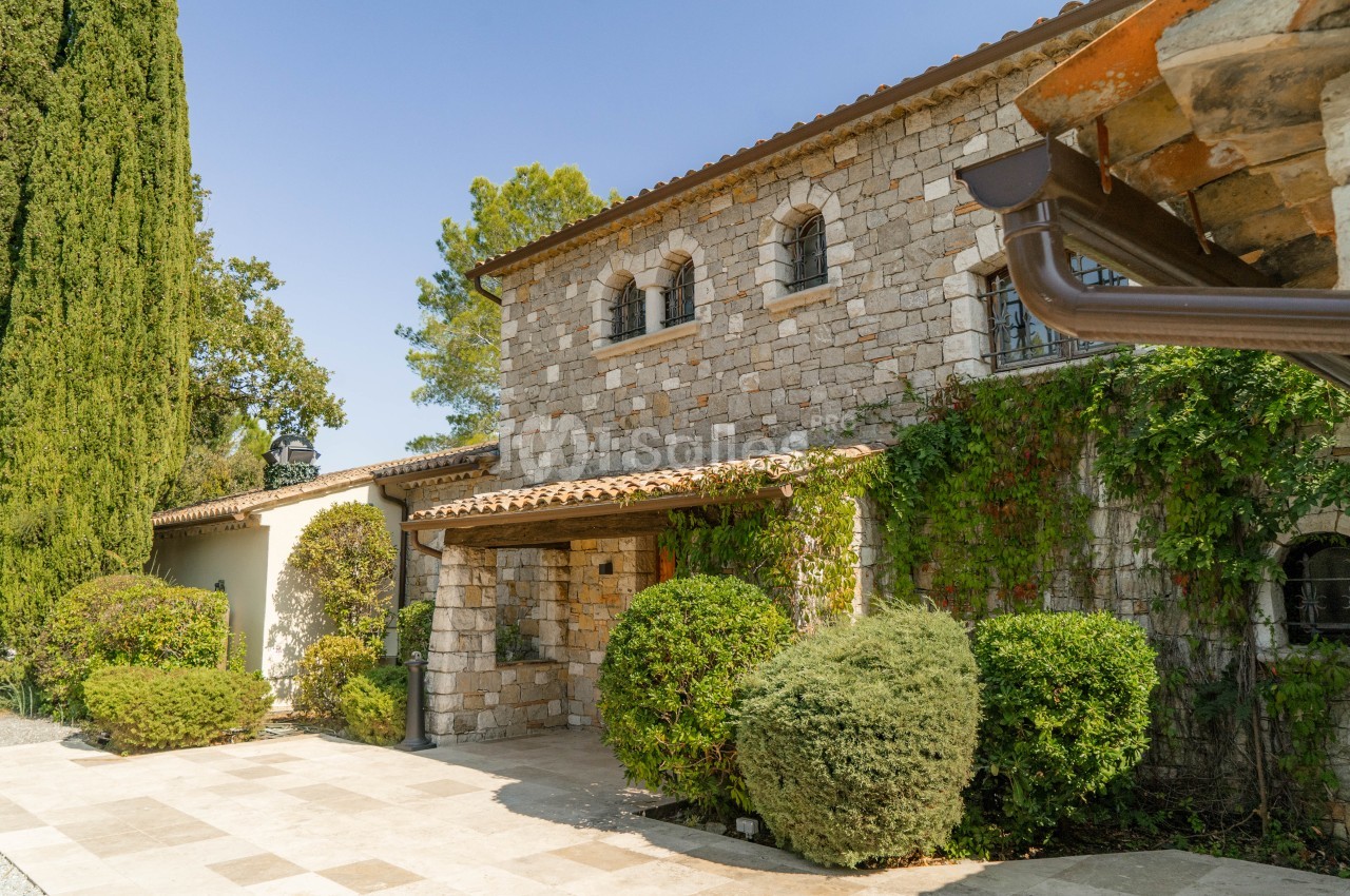 Façade en pierre d'une villa provençale avec entrée couverte, arbustes taillés et cyprès - jour ensoleillé.