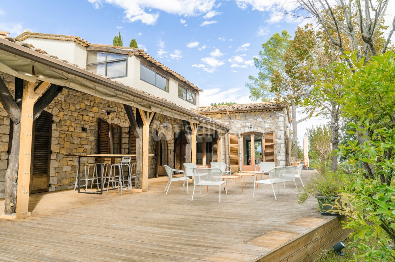 Terrasse en bois avec chaises blanches face à une maison en pierre avec volets en bois.