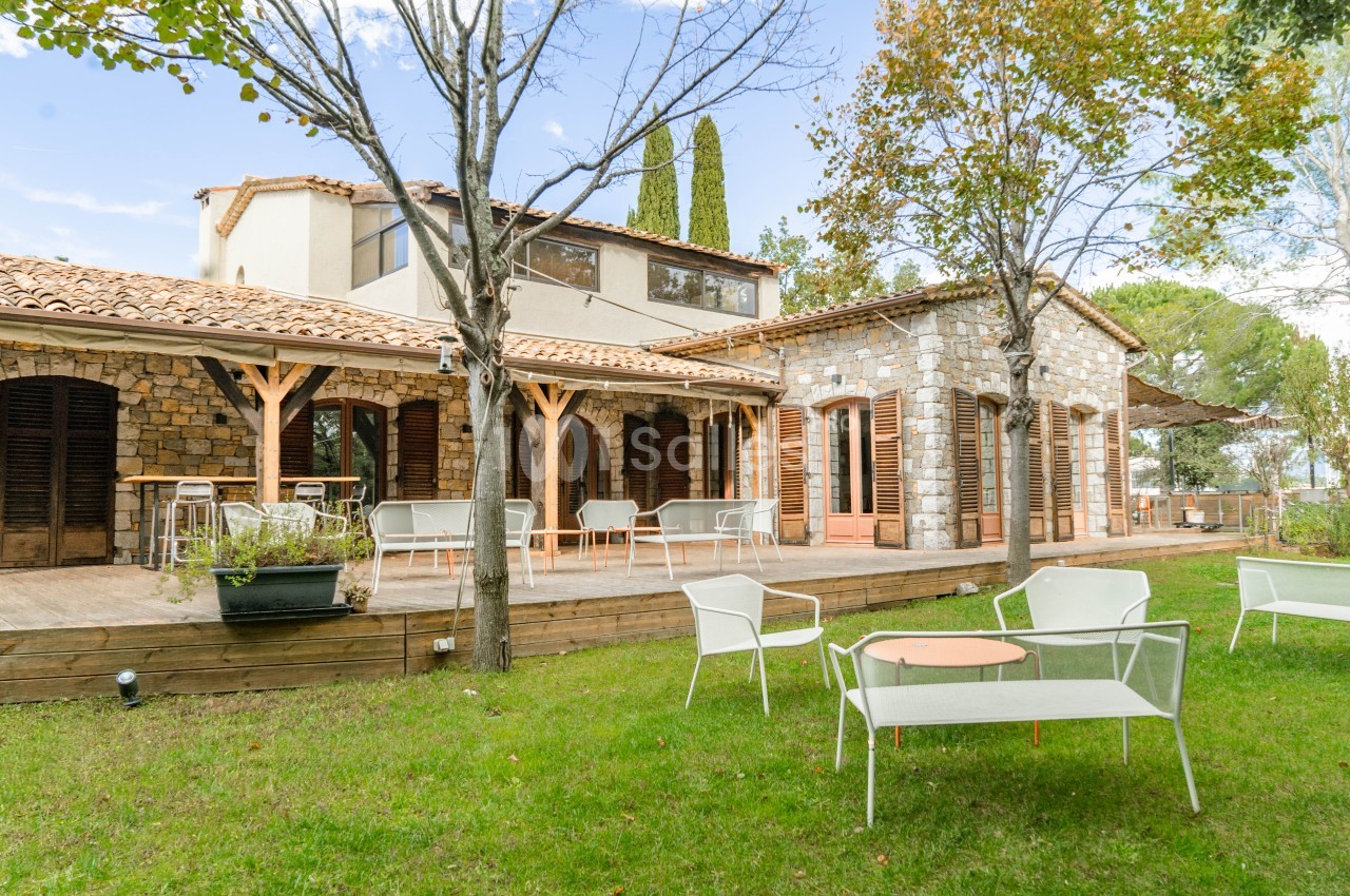 Maison en pierre avec terrasse en bois, mobilier blanc de détente et pelouse arborée.