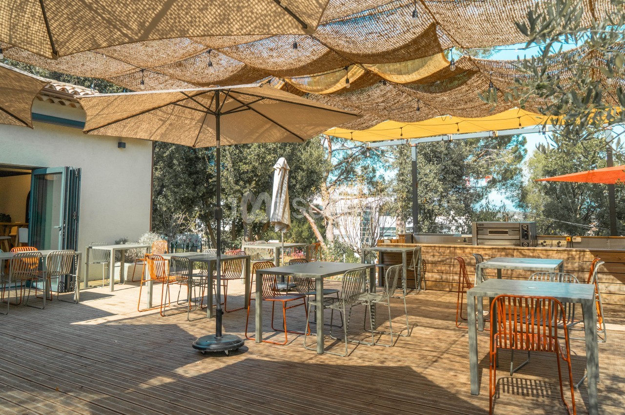 Terrasse ombragée avec tables, chaises orange et jaunes, parasols, cuisine extérieure et oliviers.