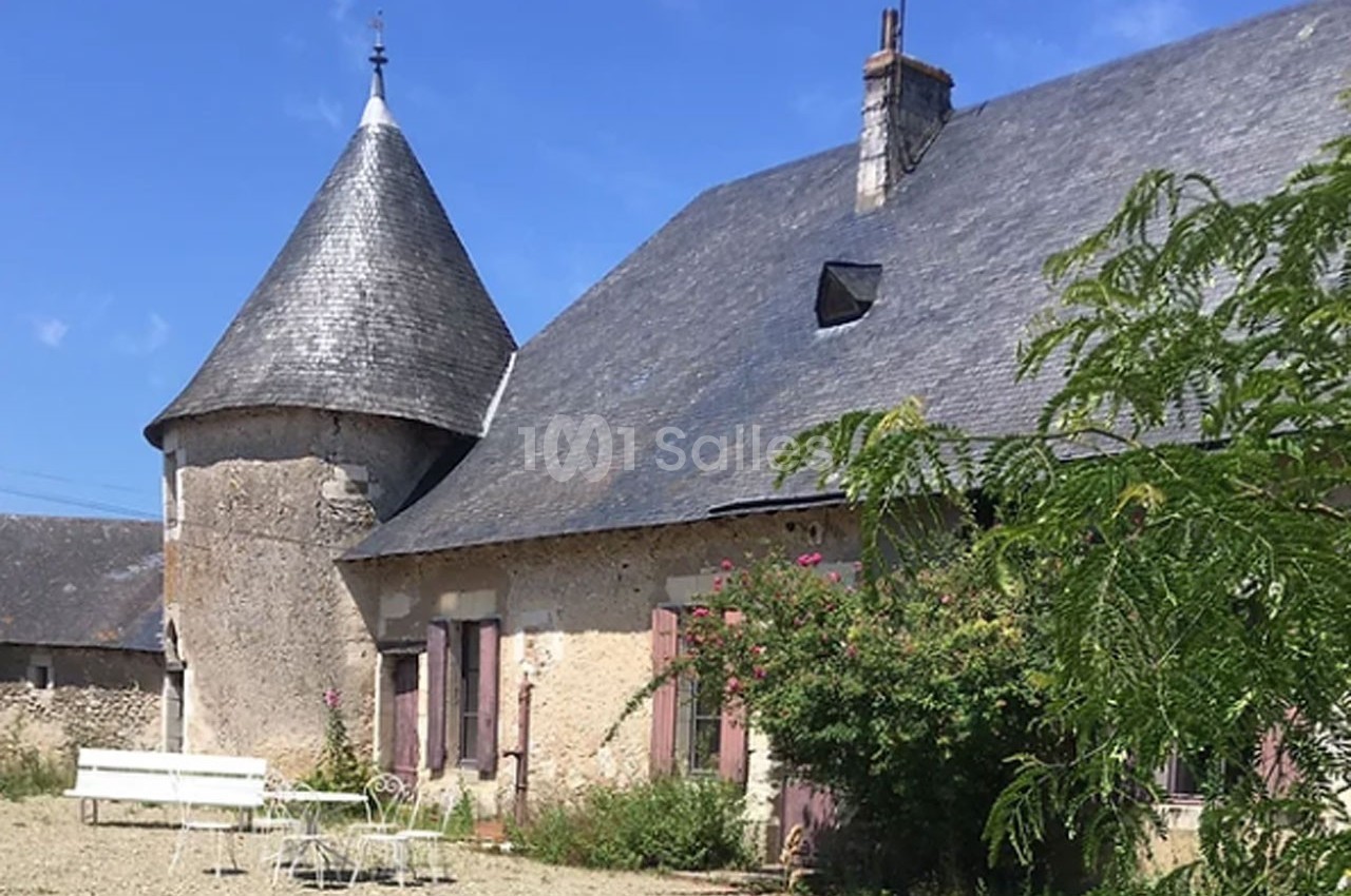 Maison en pierre avec une tour ronde, toit en ardoise, volets roses et mobilier de jardin blanc sur une cour gravillonnée.
