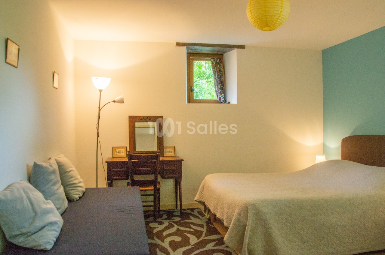 Chambre lumineuse avec un lit double, un canapé, un bureau en bois et une fenêtre donnant sur un espace vert.