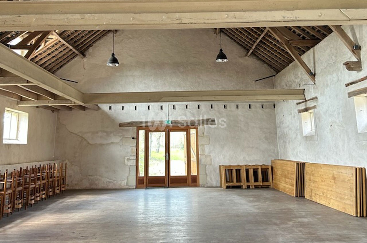 Salle vide avec murs en pierre blanche, poutres apparentes, parquet en bois et chaises empilées sur le côté.