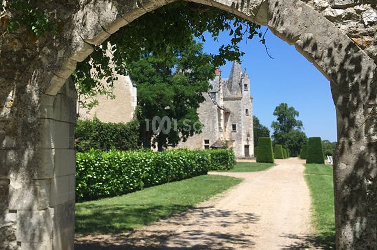 Vue d'une allée bordée de verdure menant à un bâtiment en pierre sous une arche en pierre ancienne.