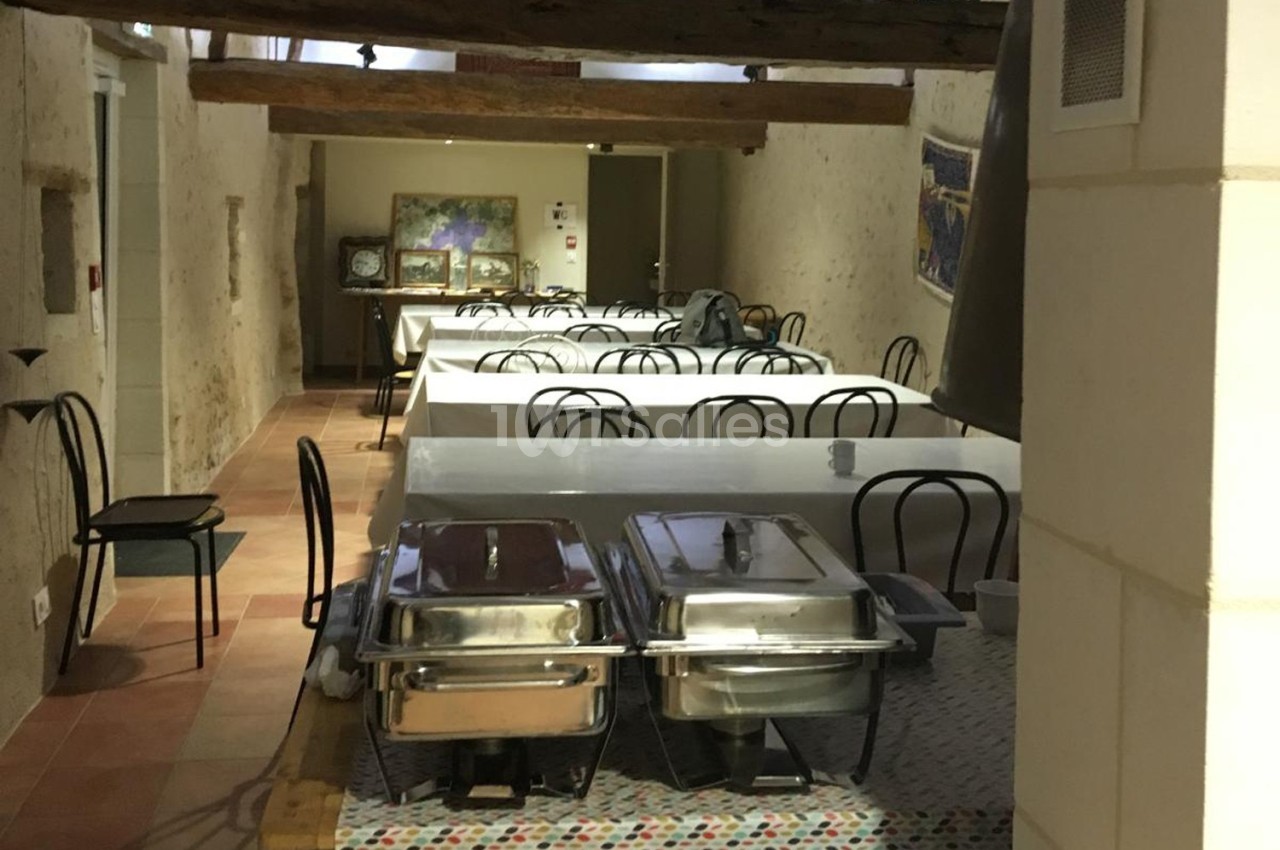 Salle de réception avec tables alignées, nappes blanches, chaises noires et plats en inox sur une table au premier plan.